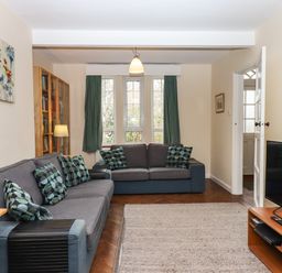 A living room with two sofas two windows with curtains a wall painting a TV on a TV stand and a bookshelf at Greenhedges in Budleigh Salterton