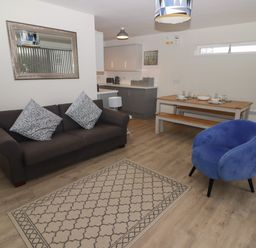 A living room with a dark sofa, blue armchair, patterned rug, dining table with bench, and kitchen area at 16 Coedrath Park in Saundersfoot