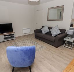 A living room with a blue chair a gray sofa with cushions a TV on a stand and a side table with a lamp at 16 Coedrath Park in Saundersfoot