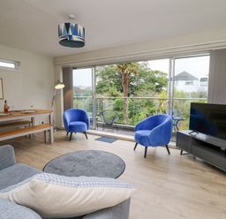 A living room with blue armchairs a sofa a TV on a cabinet a dining table and sliding glass doors at 36 Coedrath Park in Saundersfoot