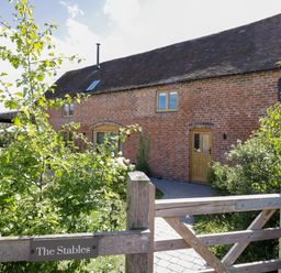A brick house with wooden doors and windows behind a wooden gate with green plants at The Stables in Droitwich Spa