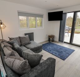 A living room with a gray sectional sofa a blue rug a wall-mounted tv and glass doors leading outside at Goldthorne Lodge in Clee Hill