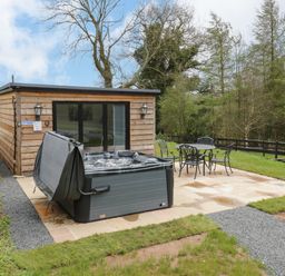 An outdoor patio with a hot tub and a metal table with four chairs next to a wooden cabin at Goldthorne Lodge in Clee Hill