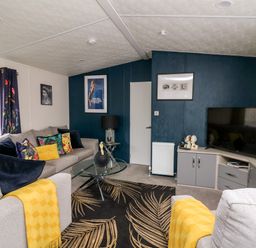 A living room with a grey sofa with colorful cushions and yellow throws a glass coffee table a black lamp a patterned rug a flat screen TV on a cabinet and framed pictures on blue and white walls at The Hideout in Gilfachrheda near Llanarth