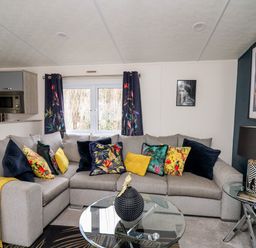A living room with a grey corner sofa with colorful cushions a glass coffee table and side table with a lamp at The Hideout in Gilfachrheda near Llanarth