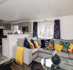 A living room with a gray sofa decorated with colorful pillows and a yellow throw next to a glass coffee table and a kitchen area at The Hideout in Gilfachrheda near Llanarth