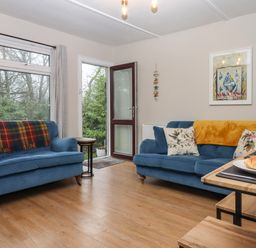 A living room with two blue sofas a wooden dining table with croissants and large windows showing trees at 2 The Glade in Kilkhampton