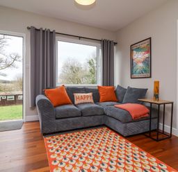 A living room with a gray sectional sofa with orange cushions and a patterned orange rug at 15 The Park in Kilkhampton