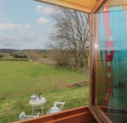 A view of a green field with trees and hills seen through an open wooden window at Harri's Hut in Bishop's Lydeard