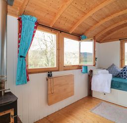 A wooden room with a stove a foldable wall table a day bed with cushions and windows with blue curtains at Harri's Hut in Bishop's Lydeard