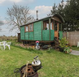 A green wooden cabin on wheels in a garden with lawn chairs a table plants and a kettle on a fire pit at Harri's Hut in Bishop's Lydeard