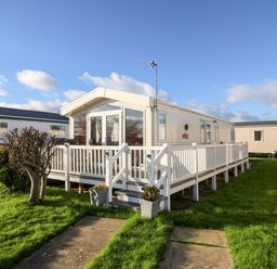 A white mobile home with a wooden porch and railing on a grassy lawn at Snape Lodge in Towyn