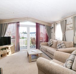 A living room with beige sofas TV wooden table and glass doors leading outside at Snape Lodge in Towyn