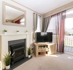 A living room with a fireplace a television on a wooden cabinet and a glass door leading outside at Snape Lodge in Towyn