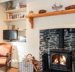 A living room corner with a wood-burning stove in a stone fireplace a wooden chair a TV on shelves and a basket of firewood at Lowen Cottage in Boscastle
