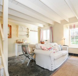 A living room with a white sofa and pillows near a window with curtains and a staircase on the left at Lowen Cottage in Boscastle