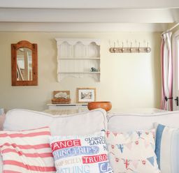 A living room with a white sofa with colorful cushions a wooden mirror a white wall shelf a coat rack and a door with curtains at Lowen Cottage in Boscastle