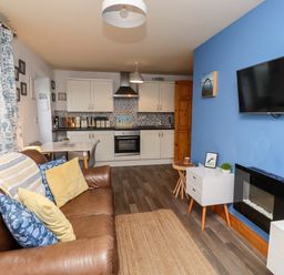 A living room with a brown leather sofa pillows a mounted flat screen TV a small white cabinet and a kitchen area with white cabinets at The Nook North Sunderland near Seahouses