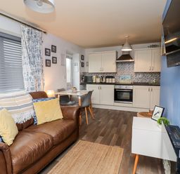 A living area with a brown leather sofa and kitchen with white cabinets and appliances at The Nook in North Sunderland near Seahouses