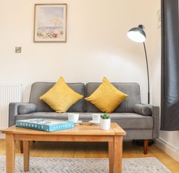 A living room with a gray sofa with yellow cushions a wooden coffee table with a Scrabble game two cups and a small plant a tall floor lamp and framed pictures on the wall at Lakeside in Falmouth