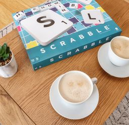 A wooden table with a Scrabble game box two cups of coffee and a small potted plant