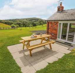 A wooden picnic table with dishes on a paved patio next to a brick building in a grassy field at The Piggery in LLanfair Talhaiarn near Abergele