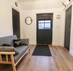 An entryway with a wooden framed gray sofa with cushions a black door a black mat on wood flooring and two black doors on the sides at Bee Cottage in Ottinge near Lyminge
