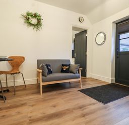 A room with a small grey sofa with patterned cushions a black door a round wall mirror a dining table with two wicker chairs and wooden flooring at Bee Cottage in Ottinge near Lyminge