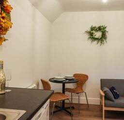 A small dining area with a round table set for two wicker chairs a gray sofa with decorative cushions and a kitchen counter with a bottle and glass at Bee Cottage Ottinge near Lyminge