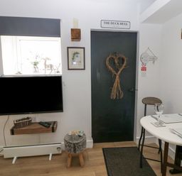 A dining area with a table set for two near a black door with a heart-shaped wreath and a wall-mounted television at High Wreah Bolt Hole in Cleator Moor