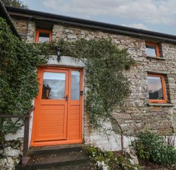An exterior view of a stone building with orange framed windows and an orange door surrounded by green plants at Ty Ceffylau in Llanllwni near Llanybydder