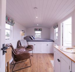 A small room with a bed elevated over cabinets two leather chairs a wooden floor a kitchenette with a sink and windows with floral curtains at Bumbles Retreat in Whepstead near Bury St Edmunds