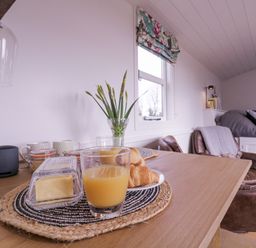 A dining table with croissants, butter, orange juice and a vase with flowers in a room with two windows at Bumbles Retreat in Whepstead near Bury St Edmunds