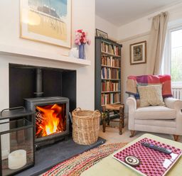 A room with a wood burning stove fire a beige armchair with a cushion and blanket a bookshelf and a window at 3 Chantry Cottages in Campsea Ashe near Woodbridge