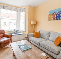A living room with a gray sofa and orange cushions a leather armchair and a wooden coffee table at The Corner Cottage in Aldeburgh