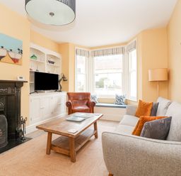 A living room with a gray sofa orange pillows a brown leather armchair wooden coffee table black fireplace and built in shelves with a TV at The Corner Cottage in Aldeburgh