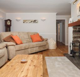 A living room with beige sofas and orange pillows a wooden coffee table a stone fireplace and a grandfather clock at Hole Park in Holsworthy