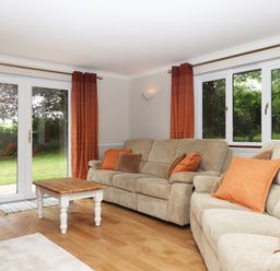 A living room with two beige sofas orange and brown cushions a wooden coffee table and glass doors with orange curtains at Hole Park in Holsworthy
