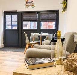 A dining area with a table set for four near black double doors and a beige sofa with a book bottle and glasses at Kingfisher Cottage in Ottinge near Lyminge