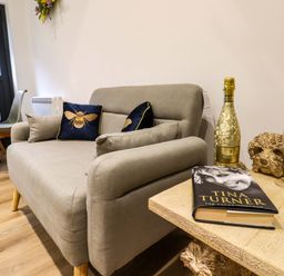 A seating area with a grey sofa, cushions, a table with a book, a bottle of prosecco and glasses at Kingfisher Cottage Ottinge near Lyminge