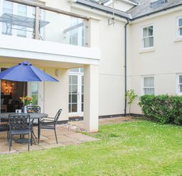 A patio area with a round table and chairs with a blue umbrella in a grassy courtyard at 5 Thurlestone Beach in Thurlestone