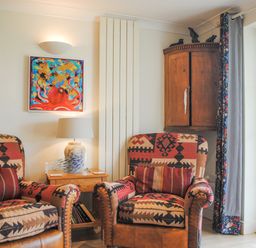 A sitting area with patterned armchairs a wooden side table with a lamp and bookshelf a wooden corner cabinet and a colorful octopus painting at 5 Thurlestone Beach in Thurlestone
