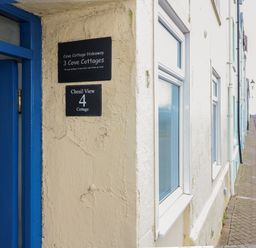 A cream colored cottage wall with a blue door and signs for Cove Cottage Hideaway and Chesil View 4 Cottage on a street with parked cars at Cove Cottage Hideaway in Chiswell On Portland