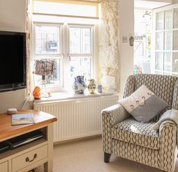 A living room with a patterned armchair by a window with vases and a small table with a television at Cove Cottage Hideaway in Chiswell On Portland