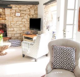 A living room with a stone fireplace armchairs a round wooden coffee table and a wall-mounted TV at Cove Cottage Hideaway in Chiswell On Portland