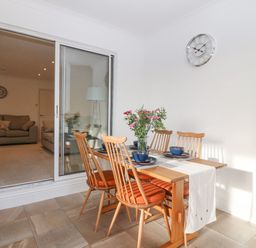 A dining area with wooden table and chairs set with blue bowls next to a sliding glass door leading to a living room with sofas at Riverview Apartment in Cemaes Bay