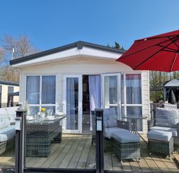 An outdoor patio with wicker furniture and a red umbrella in front of a white cabin at Ladybird Lodge South Lakes Leisure Park near Carnforth