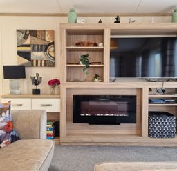 A living room with a TV and fireplace in a wooden unit next to a sofa with floral cushions at Ladybird Lodge South Lakes Leisure Park near Carnforth