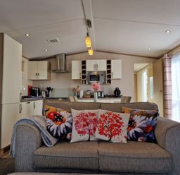 A living room with a sofa and decorative pillows facing a kitchen with cabinets and appliances at Ladybird Lodge in South Lakes Leisure Park near Carnforth