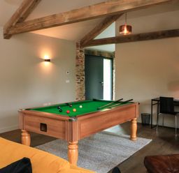 A room with a pool table on a rug next to a yellow sofa and desk with chairs at The Barns @ Downton in Dittisham